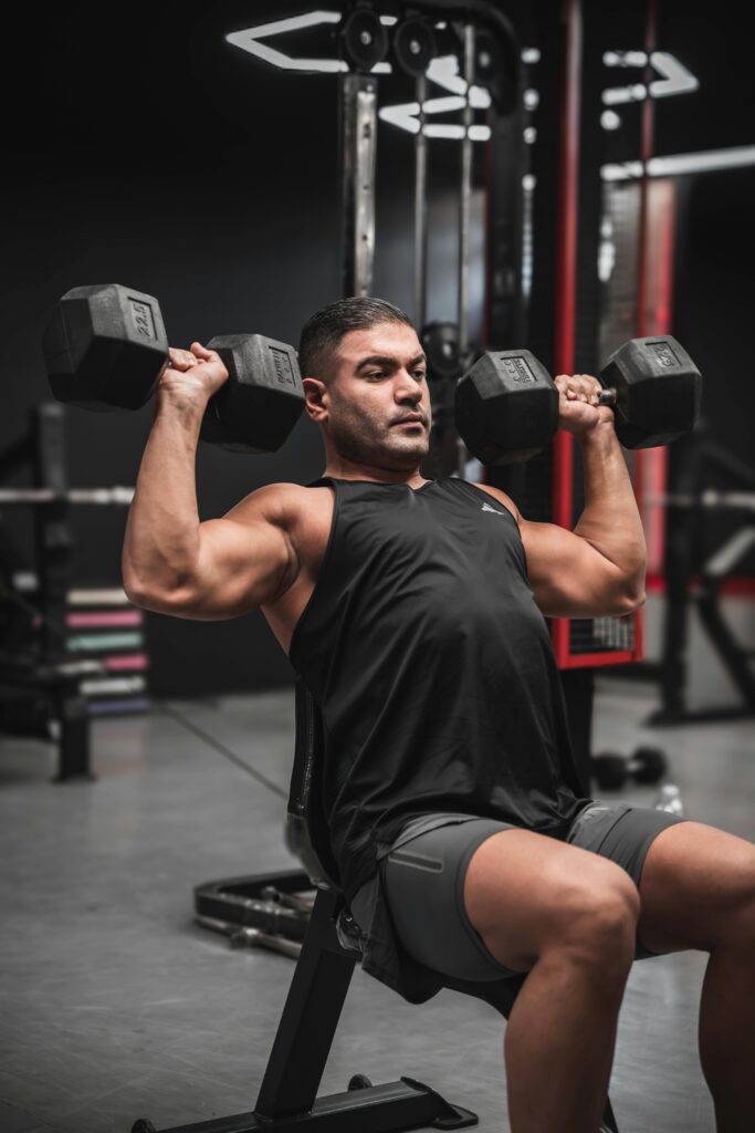 Muscular man performing shoulder press exercise with dumbbells in a gym.